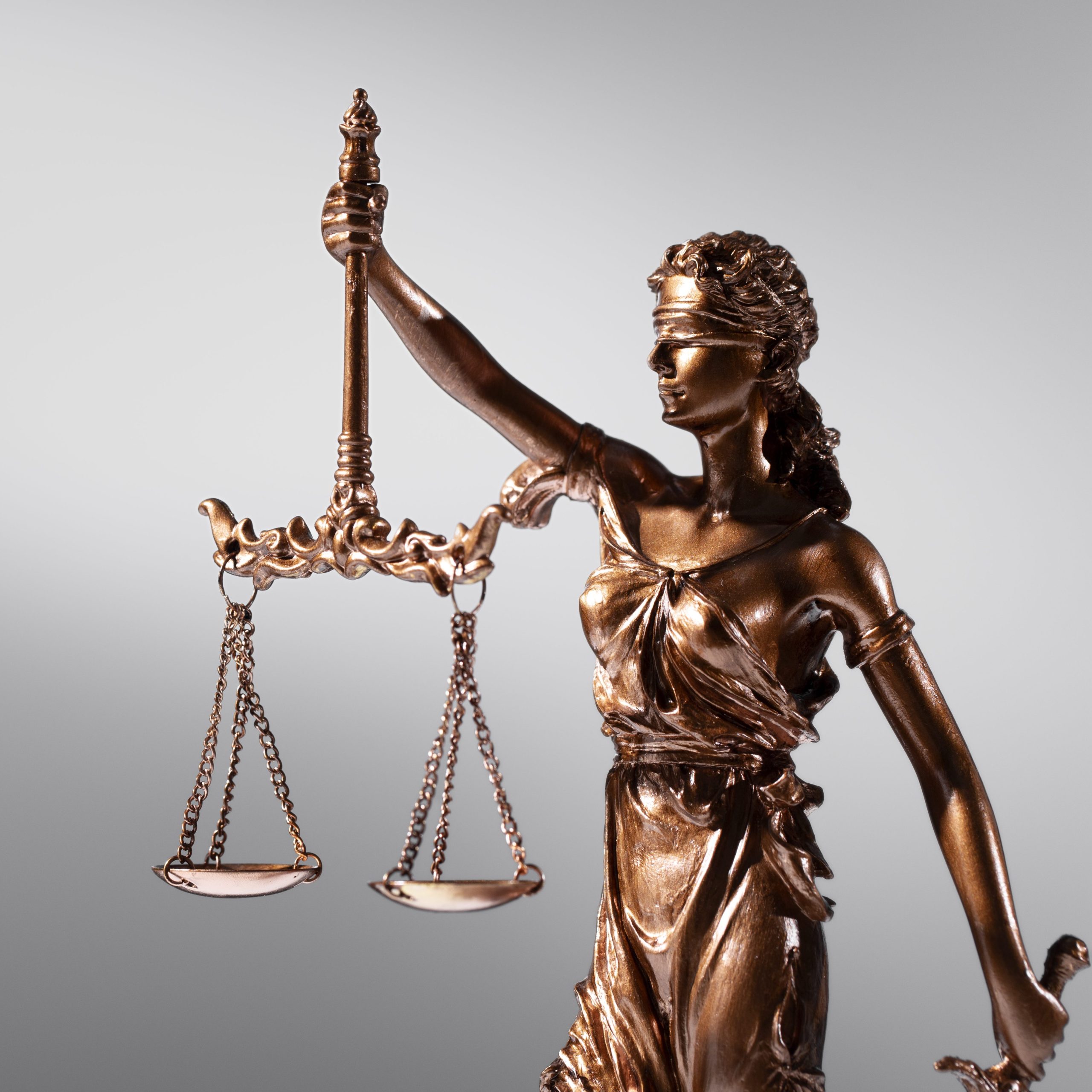 Estátua de bronze representando a Justiça, com venda nos olhos e segurando uma balança em fundo cinza claro.